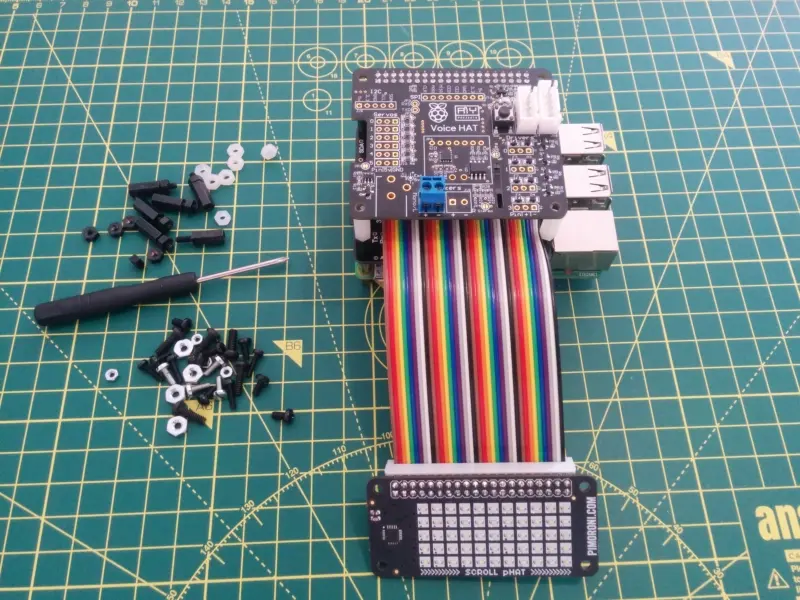 The full pHAT and multicoloured GPIO cable connected on top of the RaspberryPi with the ScrollPHAT on the end of the cable. The GoogleAIY Voice Hat sits on top of the stack.