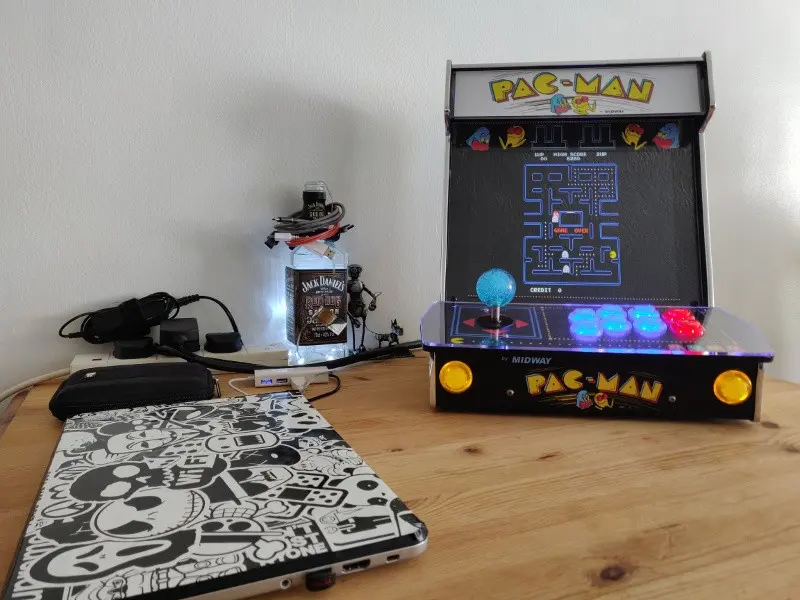 The completed cabinet shown from the front, all buttons illuminated and Pac-Man playing on the display. A laptop covered in stickers sits in the foreground.