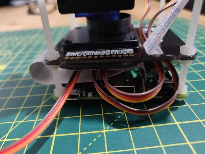 Close up of a Raspberry Pi 3A connected to a Pimoroni Pan-Tilt HAT which has a camera connect to it. The Pi has long nylon screws coming out of it. It is fully assembled and is a tight squeeze fitting all the wires between the boards.